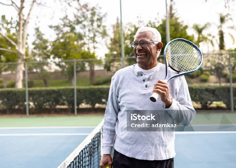 An older man laughing on a tennis court, racket in hand
